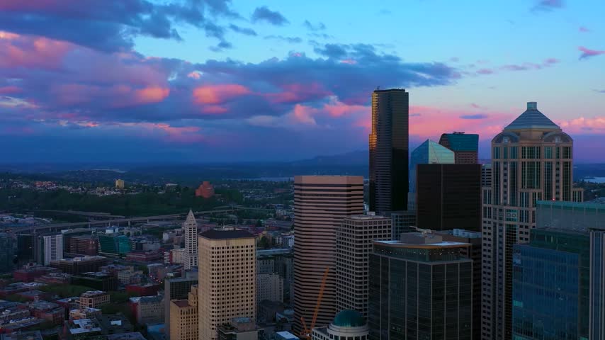 Wide high-angle panoramic aerial drone perspective looking east over the corporate skyscrapers of downtown Seattle, Washington, showcasing 1201 Third Avenue, the modern geometric glass facade of Rainier Square Tower, and a newly constructed high-rise office building set against a dramatic pink and blue sunset twilight cloud deck with distant mountain ridges.