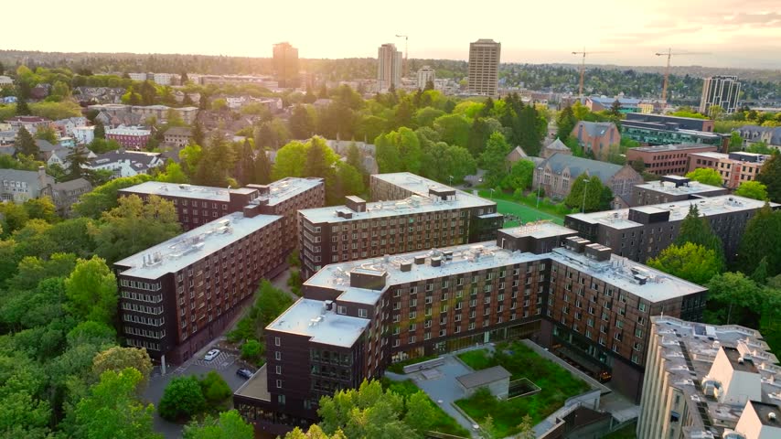 Scenic high-angle aerial drone view over the modern student residence halls on the University of Washington campus in Seattle, Washington, showcasing geometric dark brick dormitory buildings nestled within dense green tree canopies, with construction cranes and the University District skyline under a soft morning sunset cloud sky.