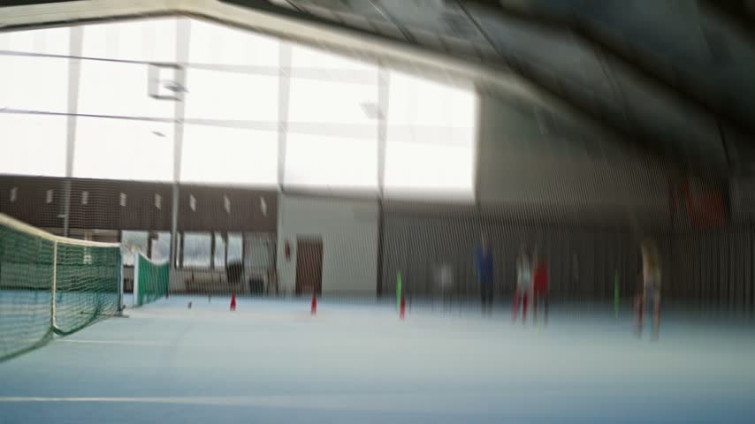 Low-angle indoor sports hall with blue floor, green divider netting, training cones, strong backlight and slight blur across spacious industrial interior, with indoor, sport, court, net detail