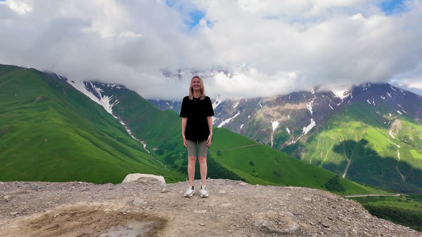 Blonde jumping with outstretched arms on top of mountain, celebrating freedom and success against backdrop of green hills, snow-capped peaks and dramatic cloudy summer skies during vacation in Georgia