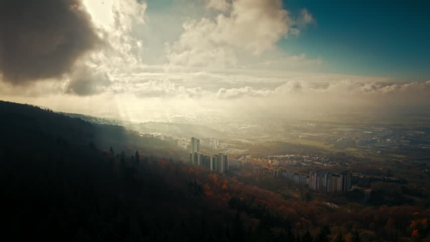 Aerial nature shot with sunbeams and layers of dramatic clouds in the sky over a European town surrounded by woodlands.