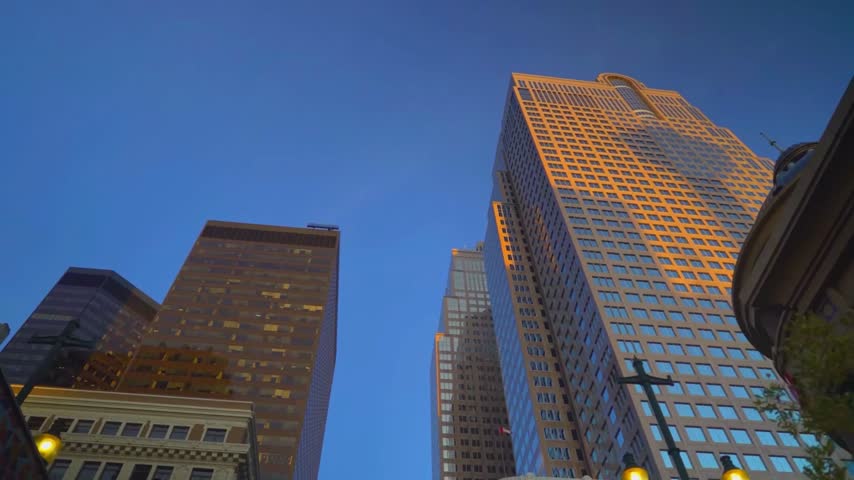 Low-angle architectural perspective looking up at the towering skyscrapers of the central business district in Calgary, Alberta, Canada, showcasing the golden stone facade of Suncor Energy Centre and neighboring glass office towers illuminated by a warm sunset glow against a deep blue evening sky.