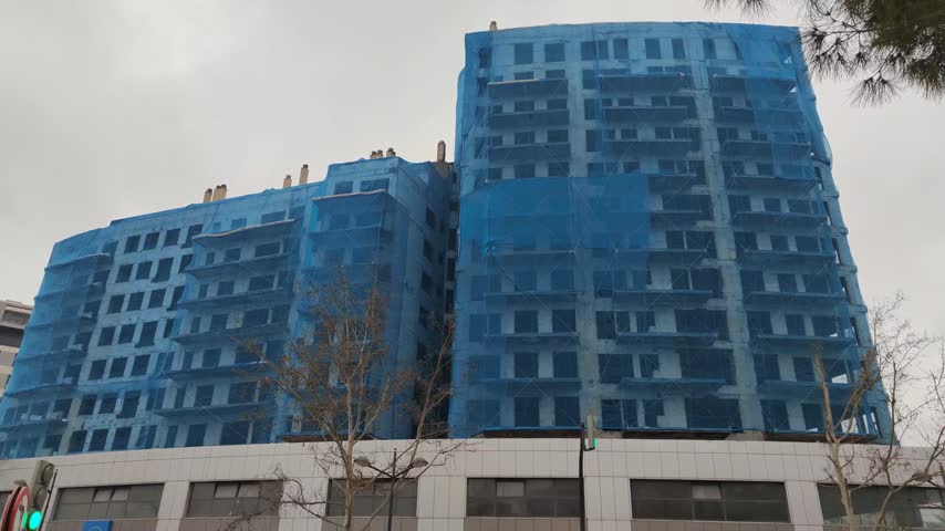 Low angle perspective of a multi story residential building undergoing construction, its facade entirely covered with blue scaffolding safety nets against a cloudy and overcast gray sky