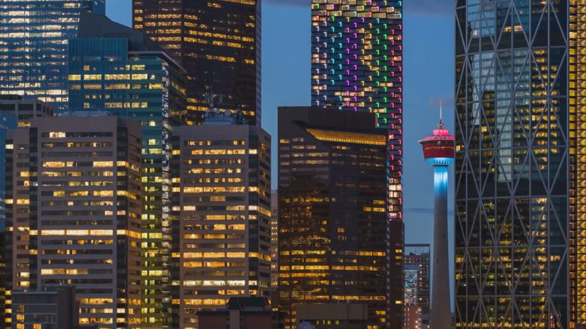 Close-up high-angle scenic view focusing on the dense skyscraper core of downtown Calgary, Alberta, Canada, at night, highlighting shifting window office lights, the moving pixelated rainbow light installation on Telus Sky, and the glowing red and blue spire of the Calgary Tower—ideal for timelapse videography transition frames.
