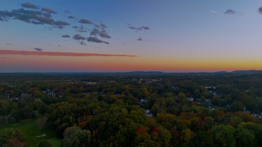 Scenic aerial view of autumn foliage, residential rooftops, and rolling hills beneath a pastel sunset sky, capturing the beauty of fall in Connecticut.
