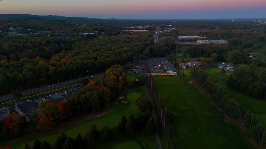 Aerial view of a golf course, winding road, parking lot, and colorful autumn forest in New Britain, CT, at dusk, showcasing suburban scenery, seasonal foliage, recreation, and local infrastructure.
