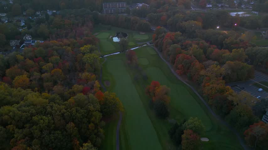 Aerial view of a golf course, winding road, parking area, and vibrant autumn forest in New Britain, Connecticut, at dusk, highlighting seasonal colors, suburban development, and outdoor recreation.