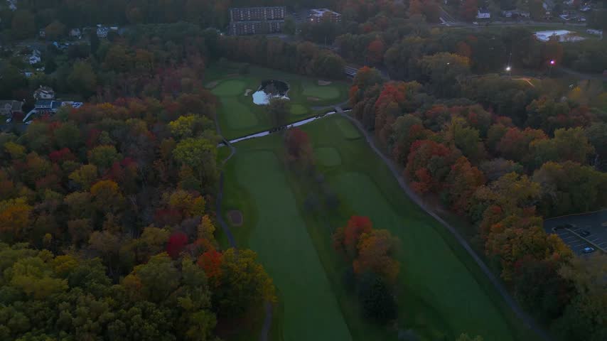 Overhead view of a golf course, road network, parking lot, and autumn woodland in New Britain, Connecticut, during evening light, showcasing fall colors, recreational spaces, and suburban landscapes.