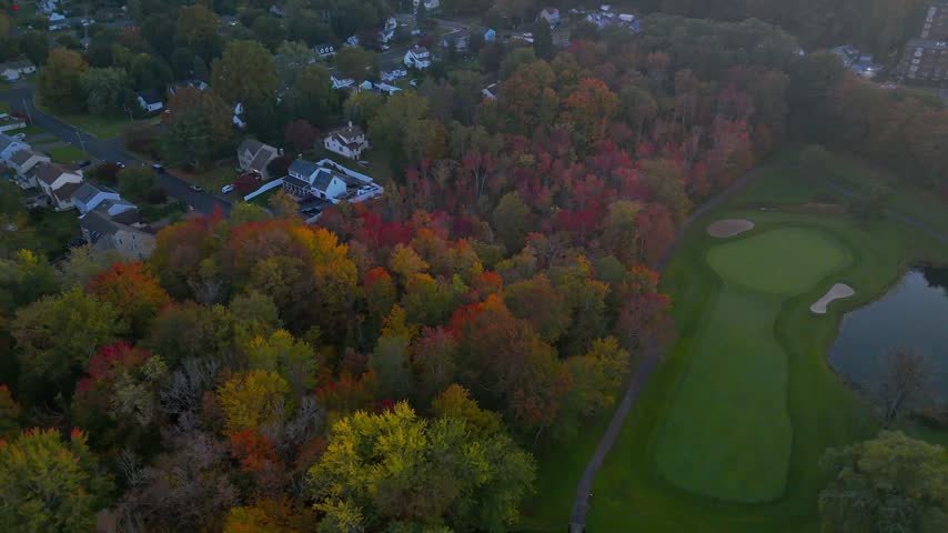 Drone footage of a golf course surrounded by vibrant autumn foliage, featuring a winding road, parking area, and nearby residential neighborhoods in New Britain, Connecticut.