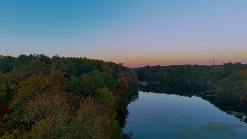 Scenic autumn fall landscape in Connecticut, USA, featuring a calm lake reflecting the sunset sky, surrounded by colorful forest foliage during magic hour.