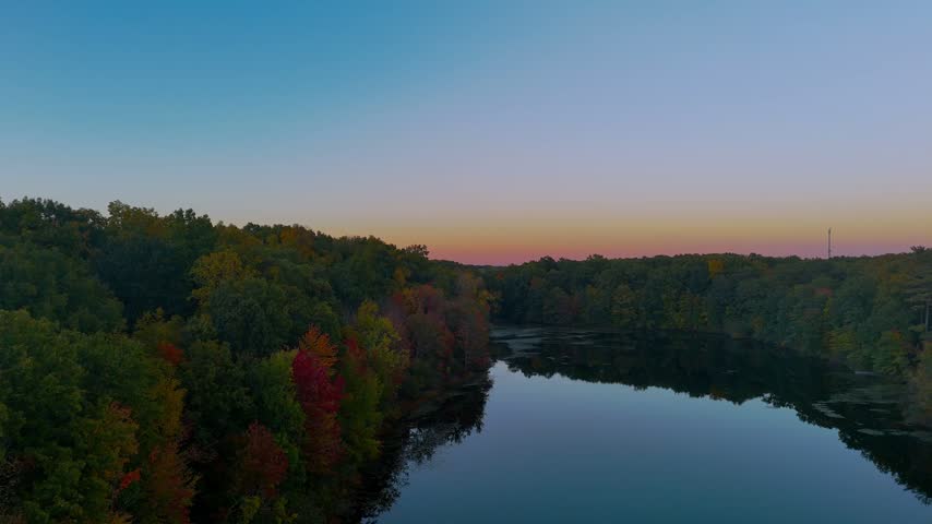 Autumn fall landscape in Connecticut, USA, featuring a calm lake reflecting the sunset sky, surrounded by colorful forest foliage during magic hour.