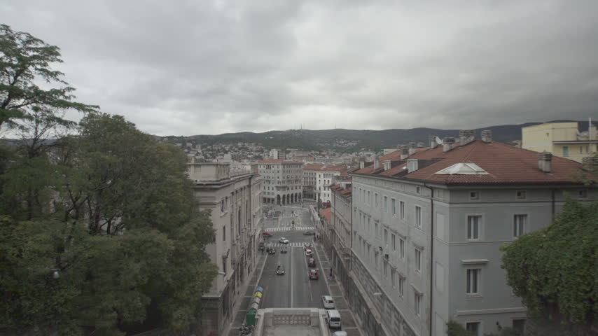 Elevated view over the centre of Trieste, Friuli Venezia Giulia, Italy, with red tiled rooftops, a street with traffic leading to a square, trees and green hills dotted with houses under a cloudy sky.