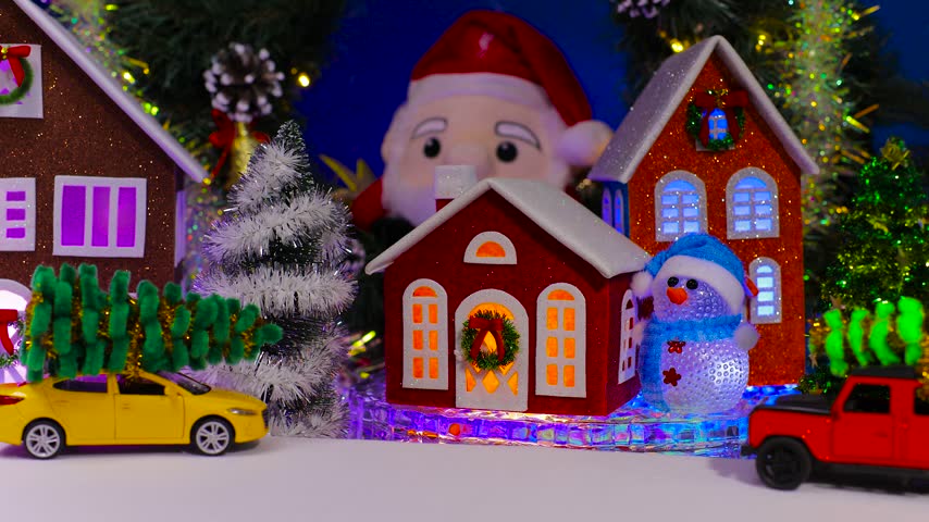 An unusual Santa Claus is riding against the backdrop of a small house with orange light in the windows and colorful toy cars nearby
