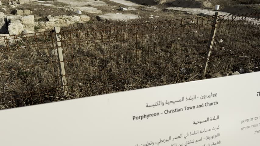 Information board overlooking the ancient stone ruins of Tel Shikmona archaeological site on the rocky Mediterranean coast of Haifa, Israel, with the modern city skyline in the background.