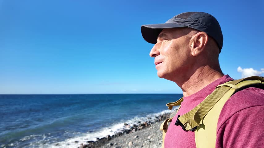 Mature man wearing a backpack and cap enjoying a sunny morning hike along a rocky beach, looking thoughtfully at the calm blue sea for mental well-being and a healthy lifestyle