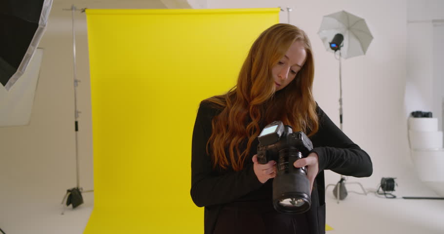 Female photographer checking DSLR settings in studio, preparing to shoot viewer on yellow backdrop. Camera, lens, flash, lighting, portrait, headshot, equipment