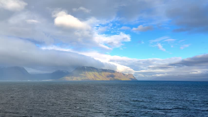 A picturesque view of the remote coastal town of Ísafjörður nestled along the dark waters of a fjord, beneath rugged, snow-dusted mountains and dramatic overcast skies.