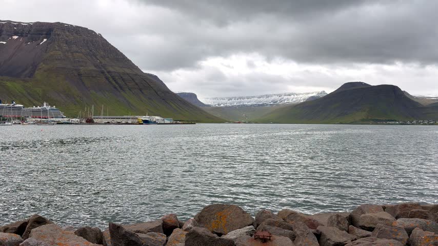 A picturesque view of the remote coastal town of Ísafjörður nestled along the dark waters of a fjord, beneath rugged, snow-dusted mountains and dramatic overcast skies.