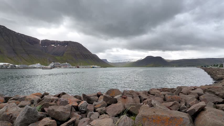 A picturesque view of the remote coastal town of Ísafjörður nestled along the dark waters of a fjord, beneath rugged, snow-dusted mountains and dramatic overcast skies.