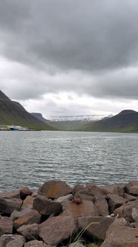 A picturesque view of the remote coastal town of Ísafjörður nestled along the dark waters of a fjord, beneath rugged, snow-dusted mountains and dramatic overcast skies.