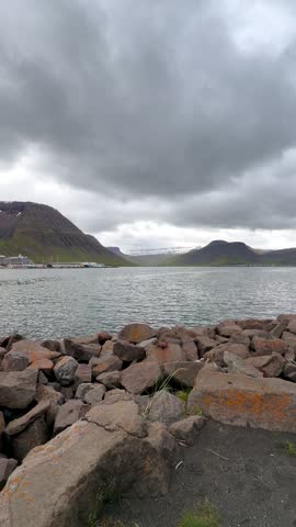 A picturesque view of the remote coastal town of Ísafjörður nestled along the dark waters of a fjord, beneath rugged, snow-dusted mountains and dramatic overcast skies.
