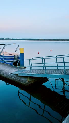 A motorized pontoon boat with a canopy is moored to a concrete dock at dusk. 