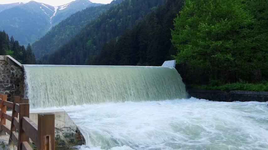 rushing water flows over a concrete dam in a scenic mountain landscape
