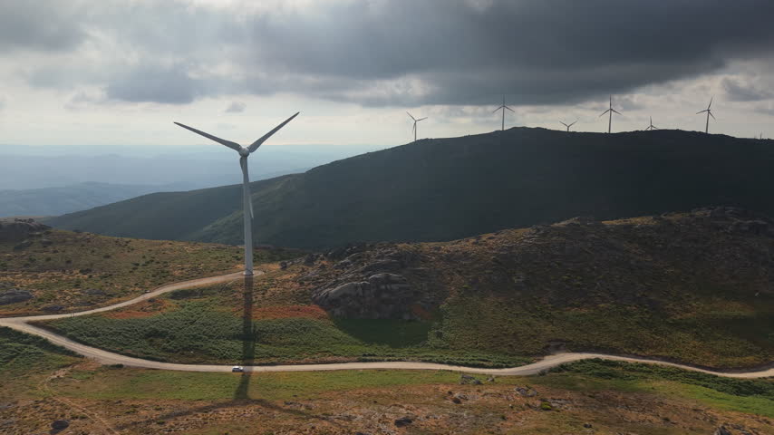 Wind turbines line a rugged mountain ridge above a winding road and green valley under dramatic clouds in Portugal.