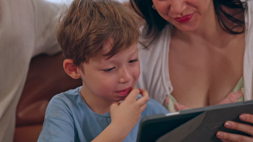Young woman and boy looking at tablet together at home, mother sharing tender family moment with her child, seated close as they enjoy quiet time and connection together