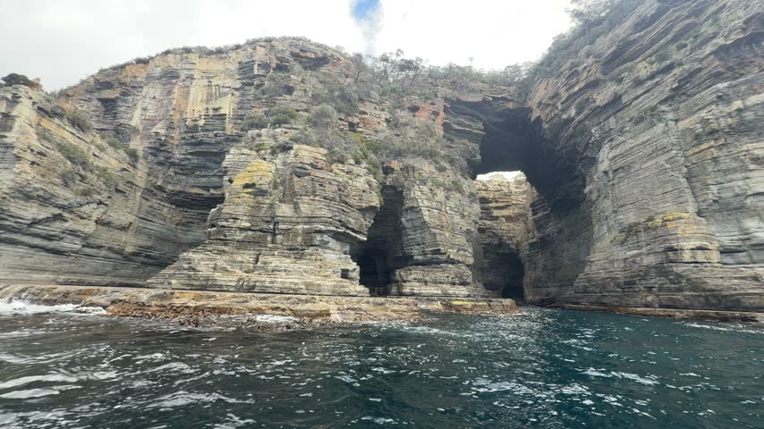 Boat approaching sea caves in layered sandstone cliffs Tasman Peninsula.