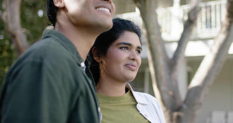 South Asian couple standing by trees and balconies, man turning to woman, smiling, sharing smiles. Duo, courtyard, greenery, daylight, eyecontact, affection, warmth