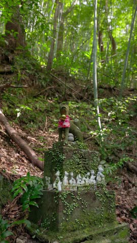 Static vertical shot of a stone base lined with offered fox figurines and a statue in a red bib.