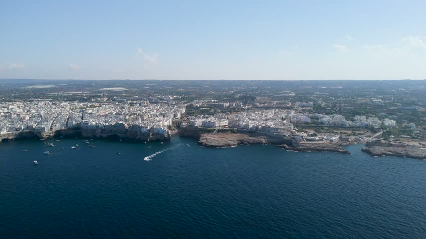 Approaching the coastal town of Polignano a Mare from the open sea, dark blue water filling the foreground and white buildings lining low cliffs on the horizon, a few boats near shore, concept of arrival and sea travel