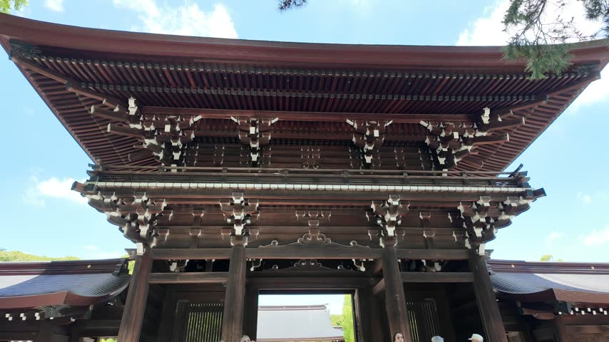 Slow push-in low angle shot of a wooden shrine gate with tiled roof, brackets and carvings against blue sky at Meiji Shrine, Tokyo Japan