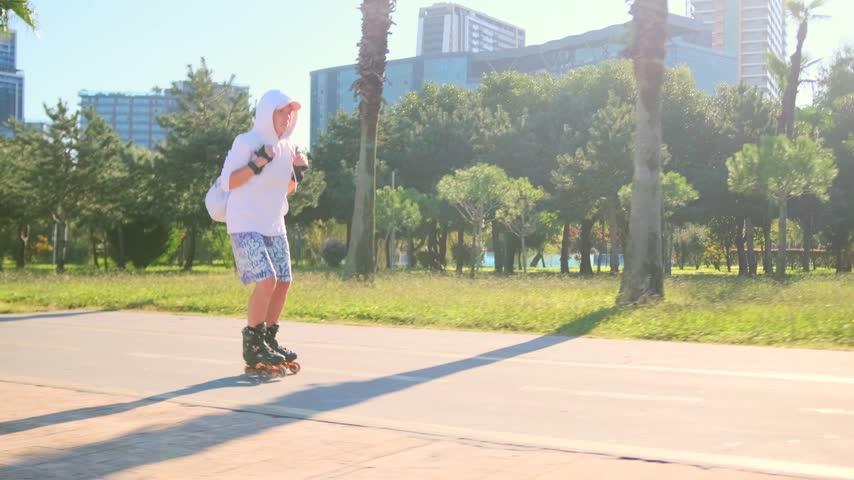 Woman explores Batumi park on inline skates, passing bench beneath palms beside tall buildings. Side full tracking shot follows rolling movement along promenade. Backpack catches sunlight against greenery.