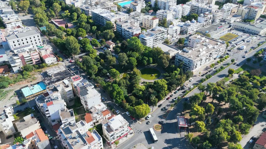 Wide drone panorama of Kalamata in Messinia, Greece, showing densely packed apartment buildings, city streets and the tree-lined Municipal Railway Park. Outdoor sports courts and a swimming pool are visible among the urban blocks, while the green slopes of the Taygetos Mountains rise in the background beneath a clear blue sky.