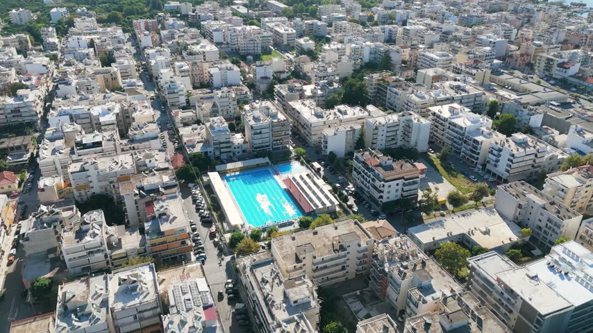 A sweeping drone perspective over the residential districts of Kalamata in southern Greece. White and beige apartment blocks form a compact urban grid, with balconies, rooftop equipment and rows of parked cars visible below. A bright blue swimming complex stands out among the buildings, while the tree-covered Municipal Railway Park adds a contrasting stretch of greenery. Toward the coast, the city