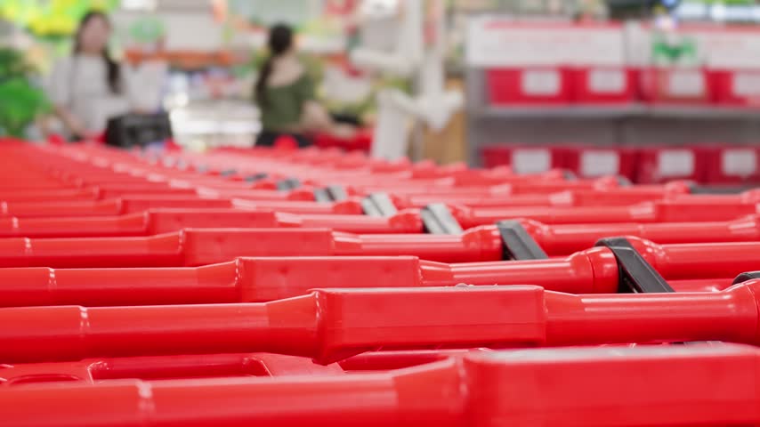 Close-up of a long row of nested red shopping carts with blurred figures of customers walking in the background inside a large modern supermarket or hypermarket. Retail trade and consumerism concept.