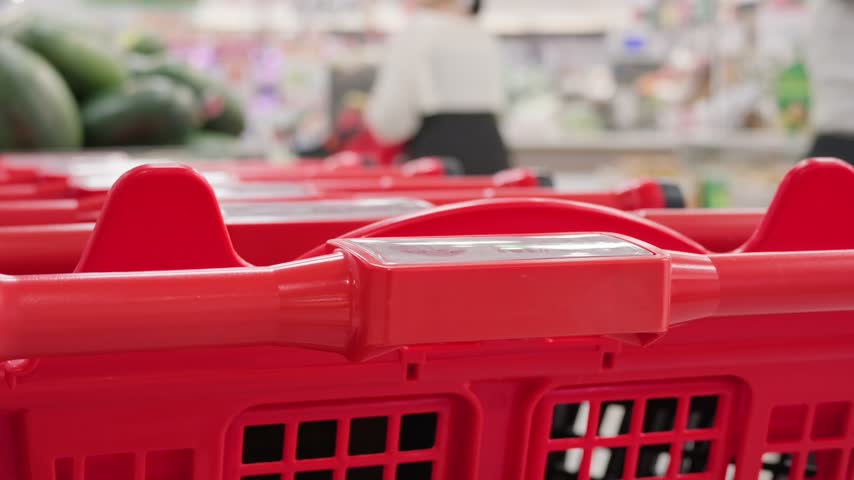 Close-up of a long row of nested red shopping carts with blurred figures of customers walking in the background inside a large modern supermarket or hypermarket. Retail trade and consumerism concept.