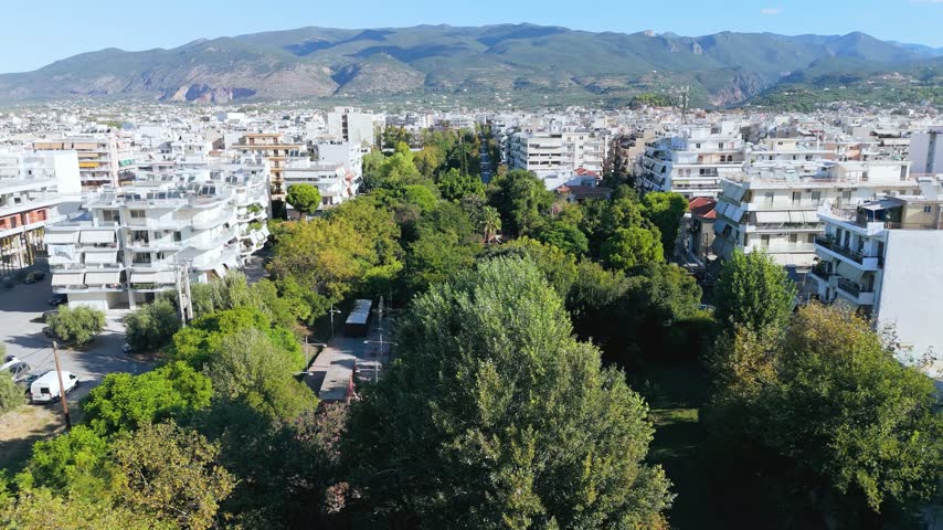 Aerial drone footage showcasing the Municipal Railway Park in Kalamata, a coastal city in the Peloponnese region of southern Greece. The camera captures a long green corridor of mature trees surrounded by densely arranged white residential apartment buildings, city streets and urban infrastructure. The impressive Taygetos mountain range forms a natural backdrop to the city. Filmed in bright daylight, the scene emphasizes the contrast between urban development and public green space. Suitable for travel documentaries, urban planning, environmental topics, Greek geography and cityscape projects.
