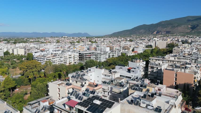 Wide aerial drone footage of Kalamata, a coastal city in Messinia, Peloponnese, Greece. The camera overlooks densely built residential neighborhoods filled with white apartment buildings, balconies and rooftops equipped with solar water heating systems and photovoltaic panels. A large green area of the Municipal Railway Park contrasts with the surrounding urban architecture. The Taygetos mountain range stretches across the background beneath a clear blue sky, highlighting the combination of modern Greek urban development, renewable energy and natural scenery.