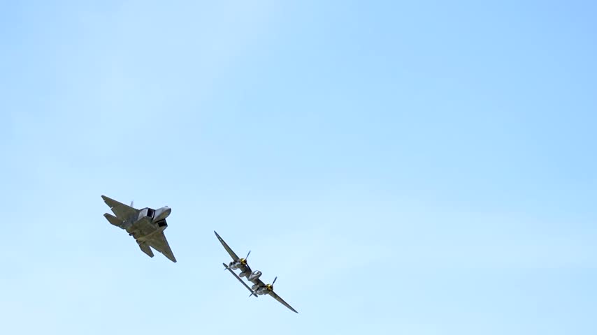 Modern stealth fighter jet banking alongside a historic twin-engine warplane during a thrilling air show demonstration in clear sky.