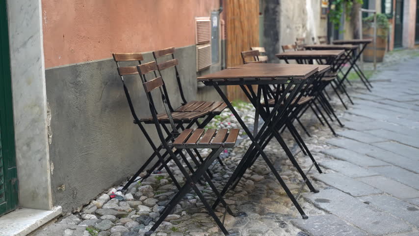 Outdoor cafe tables and folding chairs line narrow cobblestone street beside weathered plaster buildings. Empty sidewalk seating creates quiet European style setting for relaxed dining and coffee.