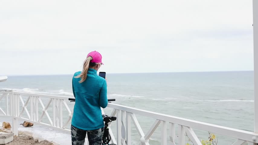 Young woman cyclist pauses beside the sea to photograph ocean waves with her smartphone.

