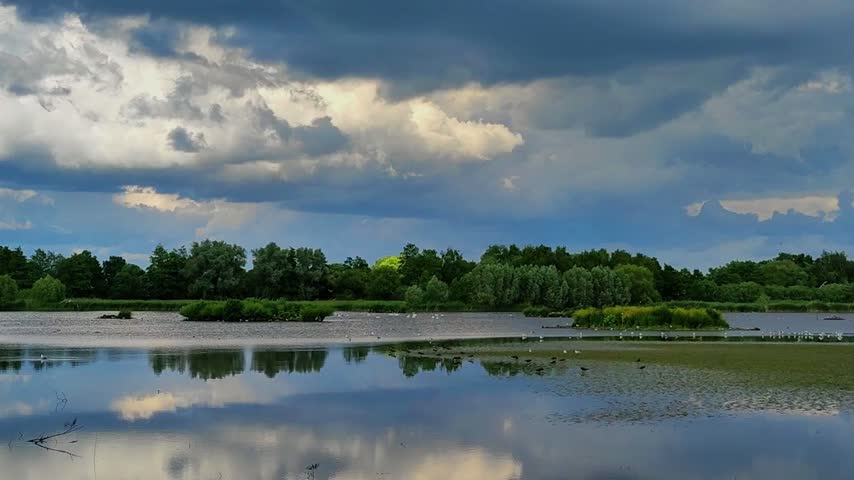Clouds move over the river in this landscape. The water reflects the sky and nearby trees. Birds can be seen near the water. It is a clear day with changing light.