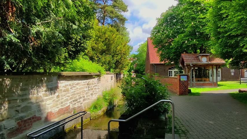 People walk near a stream and a small building surrounded by trees. The sky is clear with some clouds. The setting looks calm and natural.