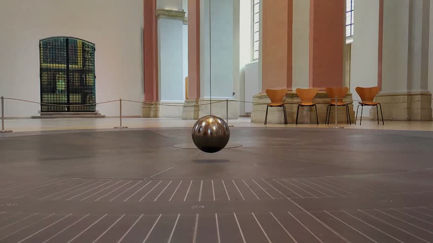 A ball moves across the floor in a museum. The large room contains wooden chairs and modern artwork. Natural light enters through tall windows, showing details of the space.