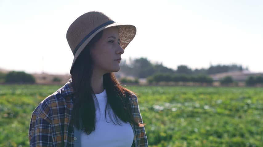 Young woman wearing a straw hat looks into the distance on a green farm field, close side profile portrait with cultivated crops and rural landscape in the background during bright daylight