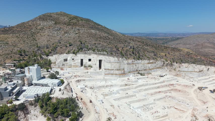 Aerial orbit shot of a marble quarry on Mount Penteli near Athens, Greece, on a sunny summer day. Dark entrances cut into the mountainside contrast with the exposed marble rock and stone debris inside the open quarry, revealing the landscape shaped by marble extraction.