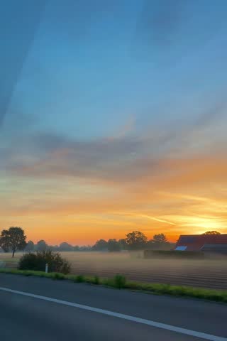 Colorful sunrise over rural fields and roadside trees viewed from a moving vehicle. Warm orange horizon beneath blue morning sky.